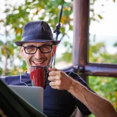 Uśmiechnięty mężczyzna w kapeluszu i okularach pije kawę, pracując na laptopie. Zastanawia się, ile można zarobić na działalności nierejestrowanej.