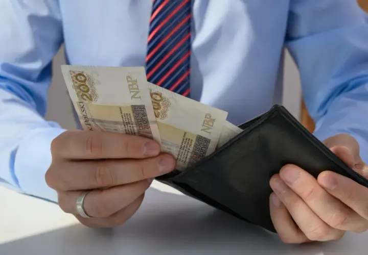 Mężczyzna w koszuli i krawacie wkłada banknoty 200 zł do portfela. Może to być wypłata, premia lub odprawa pośmiertna.