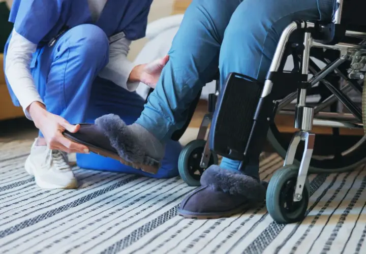 Opiekunka zakłada kapcie osobie na wózku. To codzienne świadczenie wspierające, pełne troski i empatii.