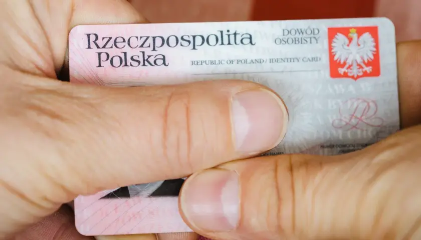 Dłonie trzymają polski dowód osobisty. Widoczna jest seria i numer dowodu, godło Polski oraz napisy "Rzeczpospolita Polska" i "Dowód osobisty".
