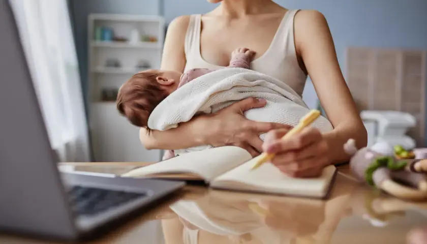 Kobieta trzyma dziecko i pisze w notatniku przy laptopie.