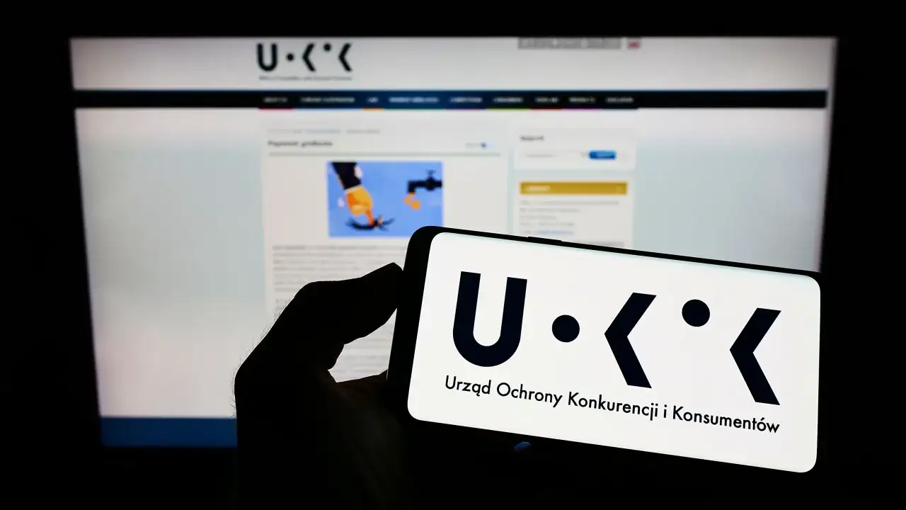 Smartfon z logo UOKiK, Urzędu Ochrony Konkurencji i Konsumentów, na tle strony internetowej. Temat: akwizycja.